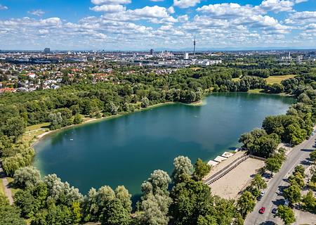 Direktlage am Lerchenauer See: 3.876 m² Ufergrundstück mit Baugenehmigung für Hotel mit Biergarten - Blick über den See mit Münchner Skyline und Alpen